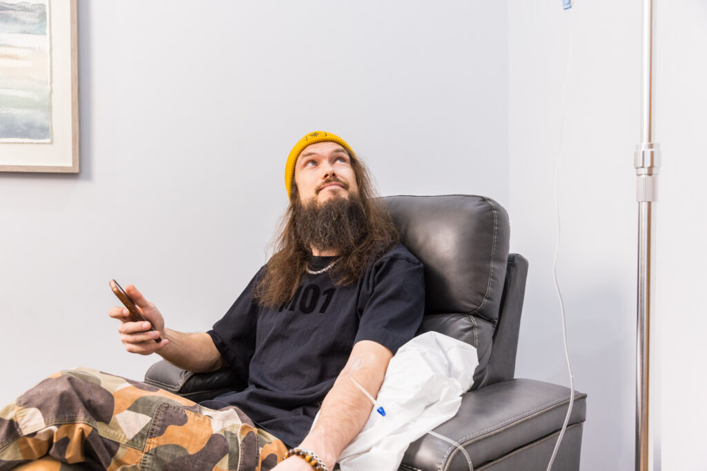 Man with long hair and a beard sits in a recliner, holding his phone while receiving an IV infusion in his arm and looking upward.