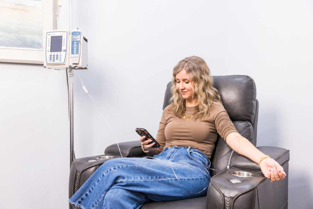 A woman sits comfortably in a recliner as she receives IV therapy and looks at her phone, reflecting on what is IV drip therapy.