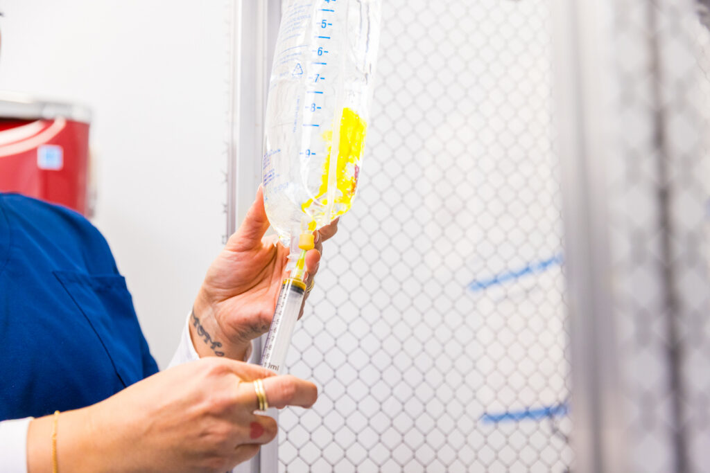 A nurse injects yellow-colored vitamins into an infusion bag filled with clear fluid.