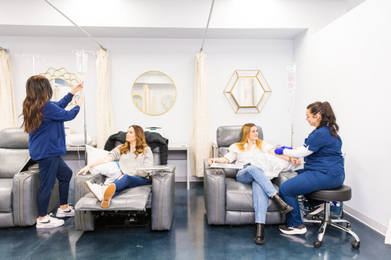 Two women sit in recliners next to each other as they receive infusions from registered nurses.