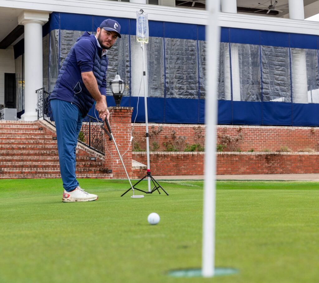 A man standing in front of a country club veranda uses his putter to practice putting while he gets his hydration therapy. He can do this because he contacted our mobile IV therapy near Mt. Juliet, TN.