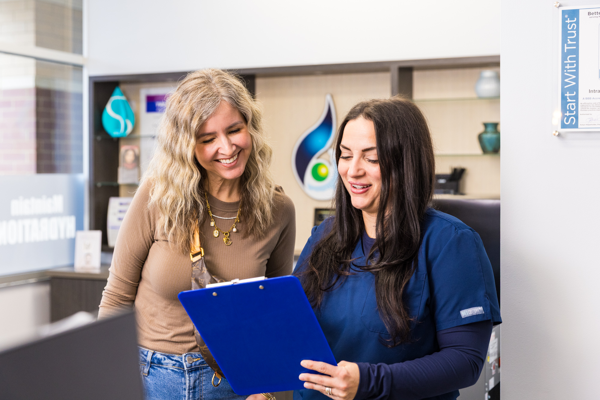 A nurse and client smile as they read papers on a clipboard in a clinic.