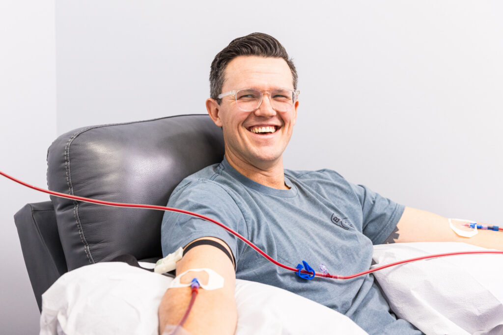 A male client smiles as he sits in a recliner and receives EBOO therapy, with both of his arms resting on cushions.
