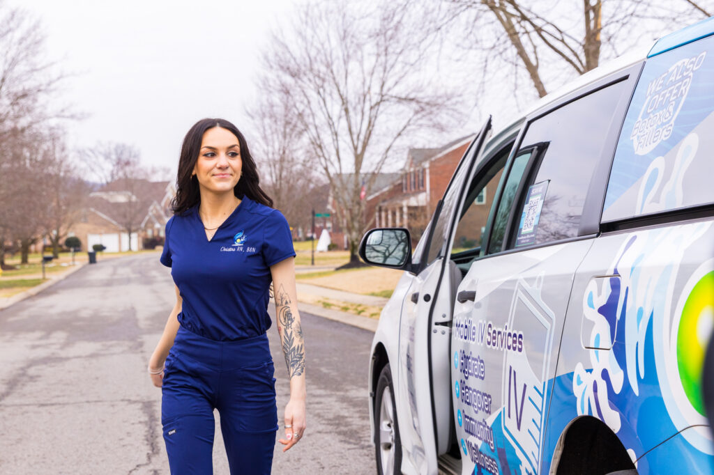 A mobile nurse in navy scrubs walking through a residential neighborhood next to a branded service vehicle to provide IV therapy at home in Nashville.