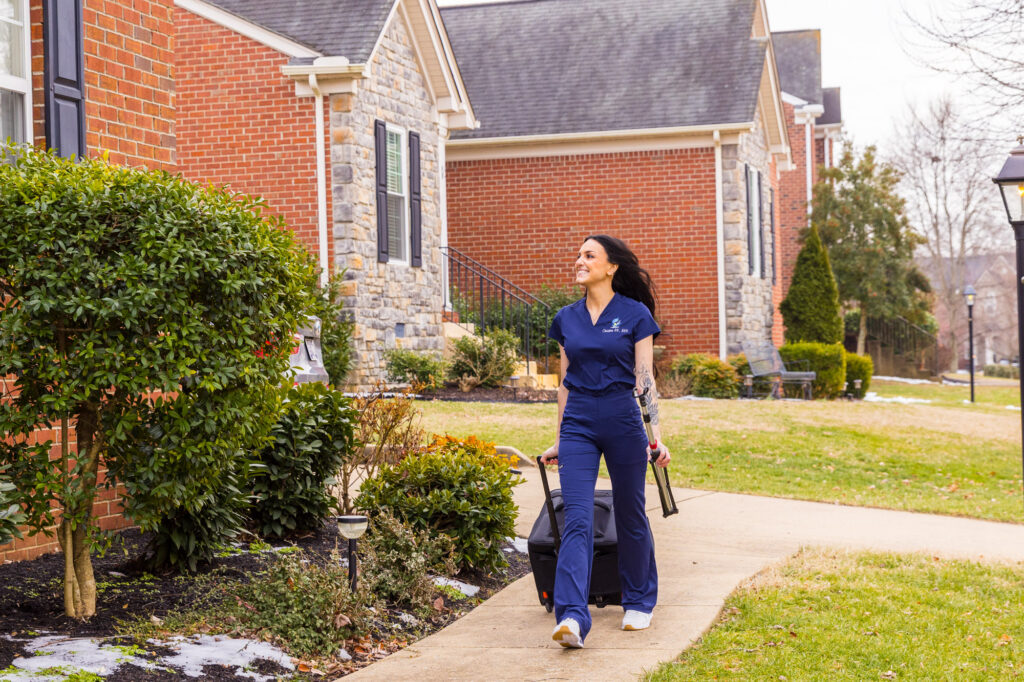 A mobile healthcare provider in navy scrubs walks toward a residential home with a medical equipment bag to provide IV therapy at home in Nashville.