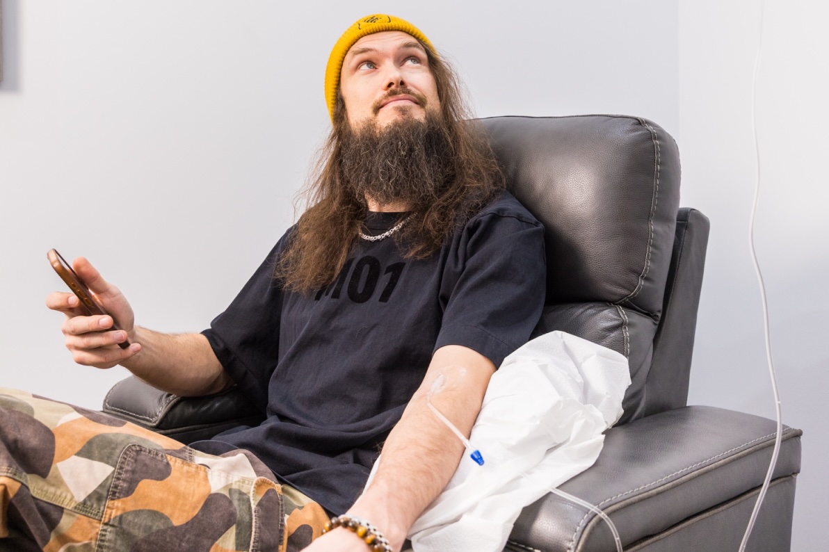 Man with long hair and a beard sits in a recliner, holding a phone while receiving an IV infusion in his arm and looking upward.